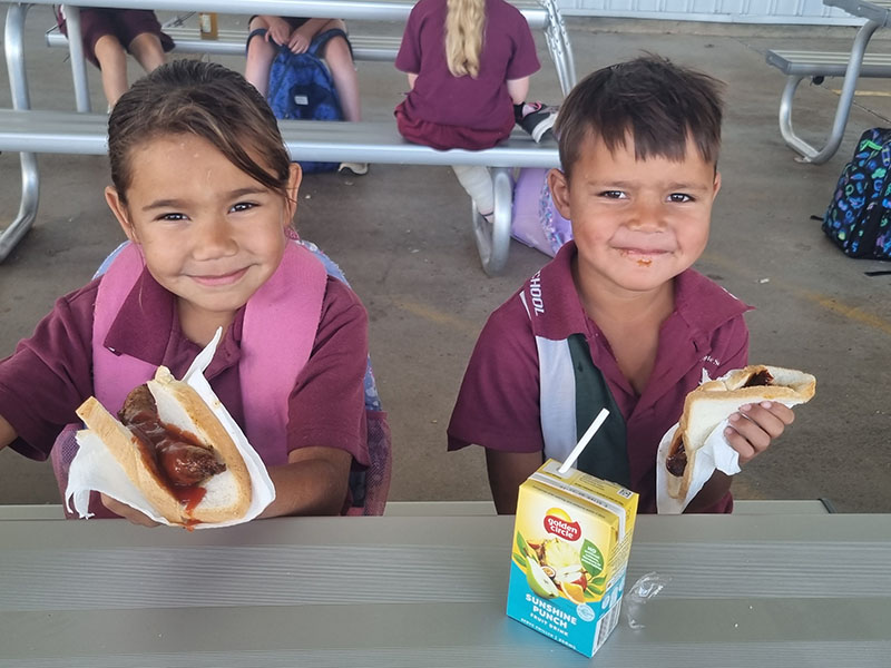 Photo of 2 happy students sitting down having sausage sizzles.