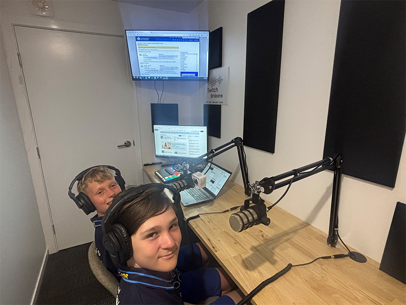 Two students in a studio sitting at a desk with headphones on and other recording equipment.