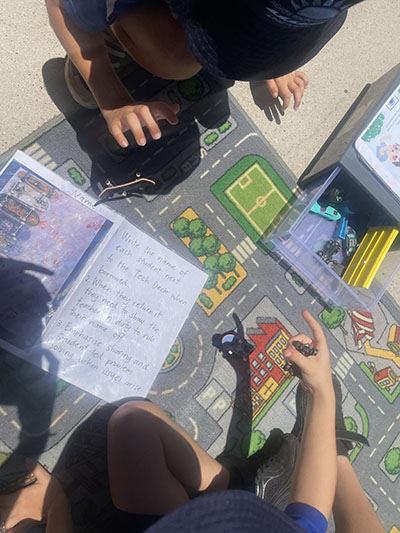 Students playing toy cars on a road map floor mat. There is an hand written instruction guide on the mat.