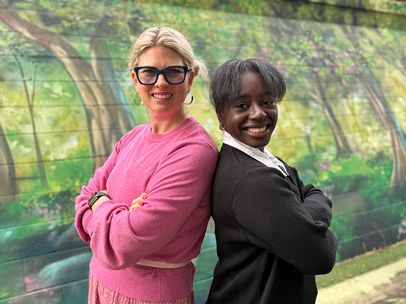 A staff member and a student are smiling at the camera, standing back-to-back.