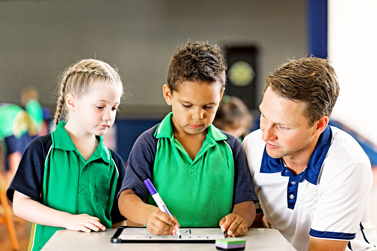 An adult and 2 children gather closely around a desk in a classroom setting, engaged in an activity together.