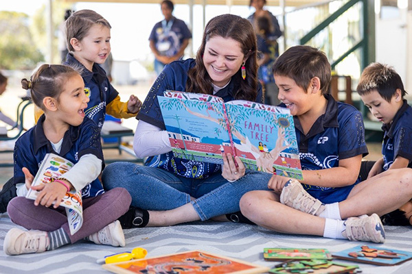 A staff is reading a book to three young children in a school setting. They are all smiling.